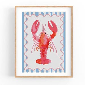 Puede incluir: Una obra de arte en punto de cruz enmarcada que presenta una langosta roja y rosa vibrante. La langosta está centrada sobre un fondo blanco, enmarcada por un borde azul y rosa. La obra se exhibe en un marco de madera.