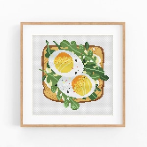 Puede incluir: Arte de punto de cruz de una rebanada de tostada cubierta con dos huevos fritos, rúcula y condimentos. La obra de arte está enmarcada en un marco de madera marrón claro. Los huevos tienen yemas de huevo de color amarillo brillante y claras de huevo blancas.