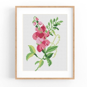 Könnte beinhalten: Ein gerahmtes Kreuzstichbild mit rosa Blumen mit grünen Blättern und Stielen. Das florale Design ist vor einem weißen Hintergrund mit einem Rastermuster angeordnet. Der Rahmen besteht aus hellbraunem Holz.
