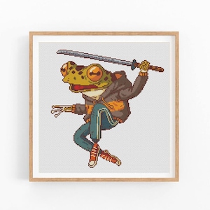 May include: A framed cross-stitch design featuring a frog character in a brown jacket, blue pants, and orange sneakers, holding a katana sword. The frog has large orange eyes and is set against a light gray background.
