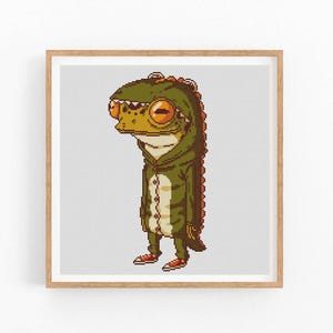 Puede incluir: Impresión artística pixelada enmarcada que presenta una rana de dibujos animados con un disfraz de dinosaurio verde y marrón. La rana lleva zapatos naranjas y tiene ojos grandes de color naranja. La obra de arte está sobre un fondo gris claro.