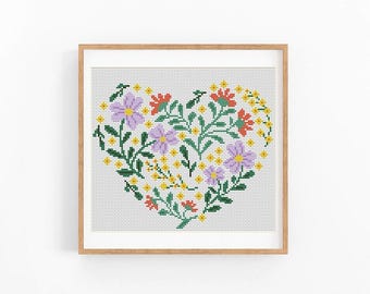 May include: Cross-stitch floral heart design in a wooden frame. The heart is filled with purple, red, and yellow flowers and green leaves. The artwork is on a white background.