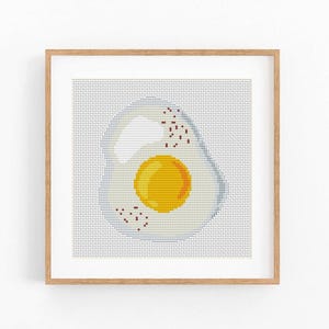 Puede incluir: Una obra de arte en punto de cruz enmarcada con un huevo frito. El huevo tiene una yema amarilla brillante y una clara de huevo blanca, con pequeños detalles marrones similares a la pimienta. La obra está sobre un fondo gris claro.