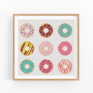 May include: A framed cross-stitch artwork featuring nine colorful donuts. Each donut has a different design, including sprinkles, chocolate, and stripes. The donuts are arranged in a 3x3 grid on a white background, framed in a light brown wooden frame.