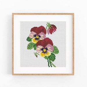 May include: A cross-stitch pattern of two pansy flowers with deep red, light purple, and yellow petals, accompanied by green leaves and stems, displayed within a square light-colored wooden frame against a plain white background.