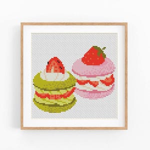 Op de afbeelding: Kruissteekkunst van twee kleurrijke macarons. Eén is groen met een rode aardbei, de andere is roze met een rode aardbei erop. Het kunstwerk is ingelijst in een lichtbruine houten lijst.