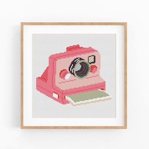 Puede incluir: Un punto de cruz enmarcado de una cámara instantánea rosa. La cámara tiene una lente negra, un botón rojo y una salida de papel verde y blanco. La obra de arte está sobre un fondo blanco y enmarcada en un marco de madera marrón claro.