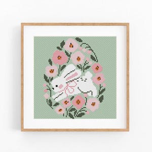 Puede incluir: Arte de punto de cruz con un conejito blanco con un lazo rosa, rodeado de flores rosas y hojas verdes, sobre un fondo verde claro. Enmarcado en un marco de madera, perfecto para la decoración de Pascua.