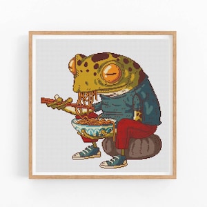 Puede incluir: Una rana verde con una camisa azul y pantalones rojos se sienta en una roca y come fideos de un tazón con palillos.