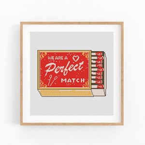 May include: A framed cross-stitch artwork depicting a matchbox. The matchbox is red and displays the words "WE ARE A Perfect MATCH" in white. The matches are visible on the right. The background is a light grey colour.
