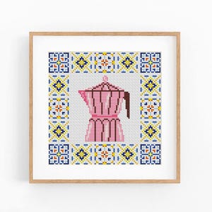 May include: A cross-stitch embroidery piece in a light wood frame featuring a pink moka pot centered within a square border of blue, yellow, and orange geometric tile patterns on a white fabric background with visible grid stitches.