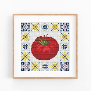 May include: A square cross-stitch embroidery featuring a vibrant red tomato with a green stem at the center, surrounded by a decorative border of blue, yellow, and white geometric tile-like patterns, all encased in a light wood frame.
