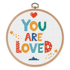 Op de afbeelding: Een witte borduurring met een kleurrijk borduurwerkontwerp met de tekst "YOU ARE LOVED". De letters zijn in verschillende kleuren en stijlen, met een rood hart bovenaan en gele sterren rond de tekst.