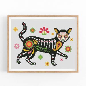 May include: A framed cross-stitch featuring a black cat decorated with Day of the Dead sugar skull makeup and floral patterns. The cat has a skeleton design and is surrounded by colorful flowers.