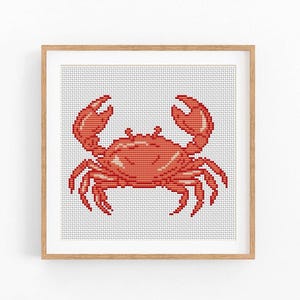 May include: A framed cross-stitch art piece featuring a red crab design. The crab is centered on a white background with a grid pattern, and the artwork is enclosed in a light brown wooden frame. The crab's claws are raised.
