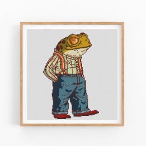 May include: A framed cross-stitch artwork of a frog character. The frog is dressed in a striped shirt, blue jeans, red braces, and red shoes. The frog has its hands in its pockets and is looking to the side. The artwork is in a wooden frame.