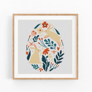 May include: Cross-stitch art in a wooden frame. The design features an egg shape filled with flowers, leaves, and two light-yellow rabbits. The color palette includes teal, orange, and cream against a white background.