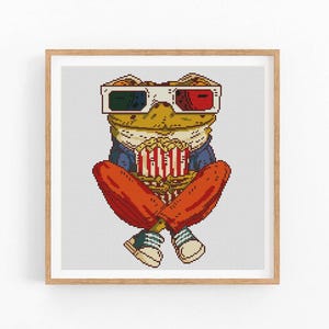 May include: A framed cross-stitch art piece featuring a frog wearing 3D glasses and holding a popcorn bucket. The frog is dressed in a blue jacket, orange pants, and sneakers. The artwork has a light background and a wooden frame.