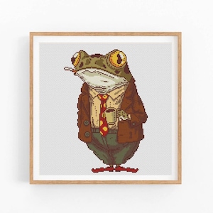 Puede incluir: Arte de punto de cruz de una rana con blazer marrón, corbata roja y pantalones verdes, sosteniendo una taza de café y un cigarrillo. La rana tiene ojos grandes y amarillos y está sobre un fondo blanco. La obra está enmarcada en un marco de madera marrón claro.