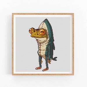 Puede incluir: Una ilustración pixelada de una rana con un disfraz de tiburón azul y gris. La rana tiene ojos grandes y redondos y una amplia sonrisa. El disfraz de tiburón tiene una aleta en la espalda y una cola.