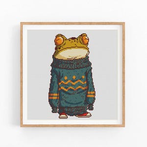 May include: A framed print featuring a cartoon frog wearing a teal sweater with orange zigzag patterns. The frog has large orange eyes and is wearing red shoes. The artwork is set against a white background with a grid pattern.