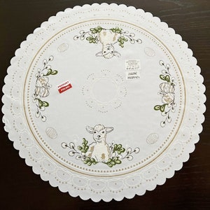 May include: White Easter-themed round tablecloth with scalloped edges. Features embroidered images of a lamb, an upside-down bunny, Easter eggs, and floral accents in green and gold. Includes text that reads "Produkt Polski".