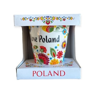 May include: A white ceramic mug with a colorful floral pattern and the text "I love Poland" on the front. The mug is in a white box with the word "POLAND" printed on the bottom.