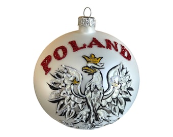 Op de afbeelding: Witte kerstbal met het woord "POLAND" in rode glitter. De bal toont een gedetailleerde illustratie van het Poolse wapen, een witte adelaar met een gouden kroon, op een witte achtergrond.