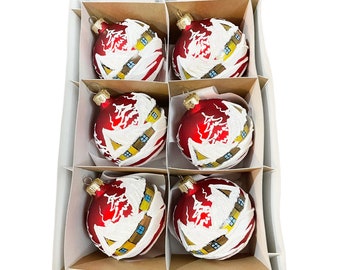 Op de afbeelding: Zes rode glazen kerstballen met witte en gouden geschilderde ontwerpen van huizen en bomen. De ballen zitten in een witte doos met bruine kartonnen scheidingswanden.