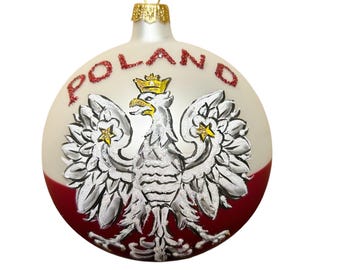 Puede incluir: Una bola de Navidad redonda con la palabra "POLAND" en purpurina roja. El adorno presenta un águila blanca con una corona dorada y una combinación de colores rojo y blanco, que representa la bandera polaca.