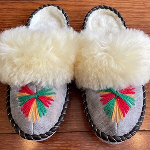 May include: A pair of gray sheepskin slippers with a black trim and a colorful embroidered design on the front. The slippers have a fluffy white sheepskin lining.