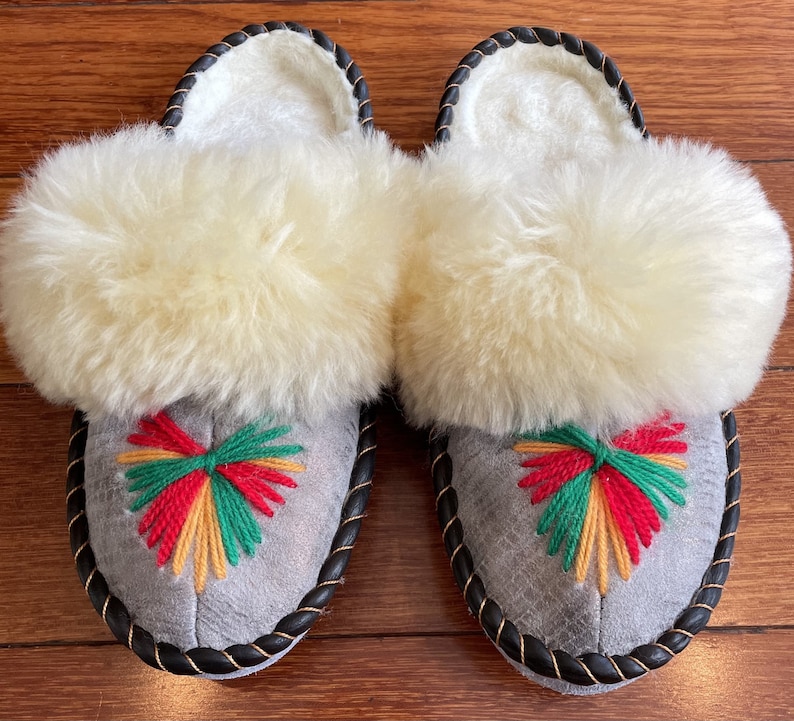 May include: A pair of gray sheepskin slippers with a black trim and a colorful embroidered design on the front. The slippers have a fluffy white sheepskin lining.