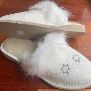 May include: White faux fur lined slippers with a white leather upper and embroidered snowflake designs. The slippers have a white sole.
