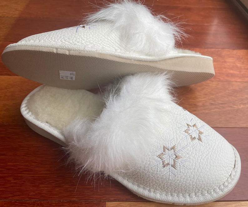 May include: White faux fur lined slippers with a white leather upper and embroidered snowflake designs. The slippers have a white sole.