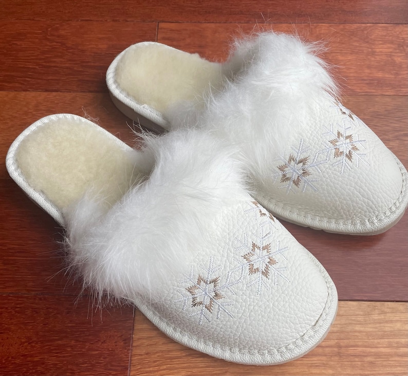 May include: Pair of white faux fur lined slippers with embroidered snowflake designs. The slippers have a white fuzzy trim around the opening.
