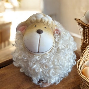 May include: A white ceramic sheep figurine with a brown face and curly white fur. The sheep has a friendly smile and is sitting on a wooden surface.