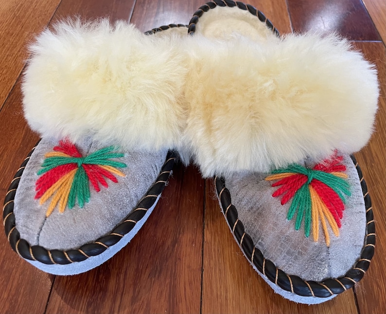 May include: Gray suede slippers with a black trim and a white fluffy lining. The slippers have a colorful embroidered design on the front.