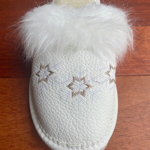 May include: White faux fur lined slippers with a white leather upper and embroidered snowflake design.