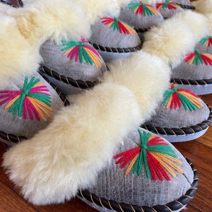 May include: A row of gray sheepskin slippers with white fur trim and colorful embroidered designs. The slippers have a black leather trim around the edges.