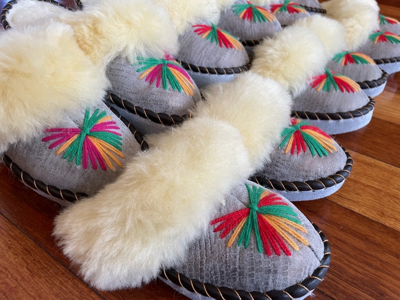 May include: A row of gray sheepskin slippers with white fur trim and colorful embroidered designs. The slippers have a black leather trim around the edges.