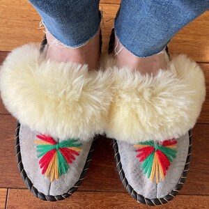 May include: A pair of gray suede slippers with white fluffy fur trim and colorful embroidered designs. The slippers have a black braided trim around the edges.