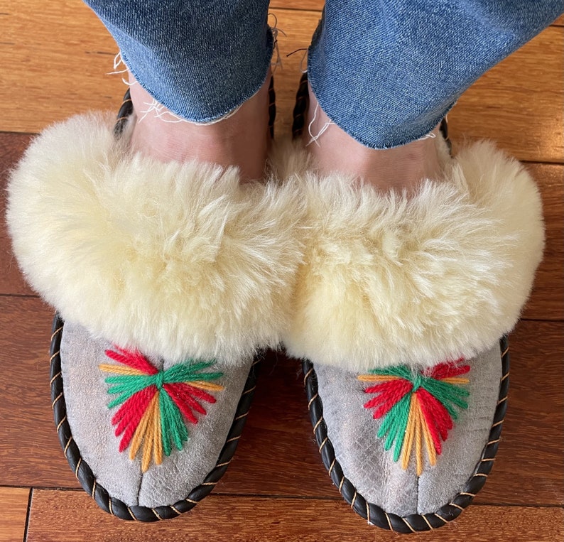 May include: A pair of gray suede slippers with white fluffy fur trim and colorful embroidered designs. The slippers have a black braided trim around the edges.