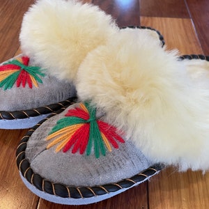 May include: A pair of gray suede slippers with white fluffy lining and colorful embroidered flowers. The slippers have a black braided trim around the edges.