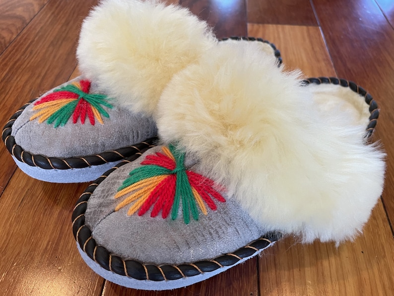 May include: A pair of gray suede slippers with white fluffy lining and colorful embroidered flowers. The slippers have a black braided trim around the edges.