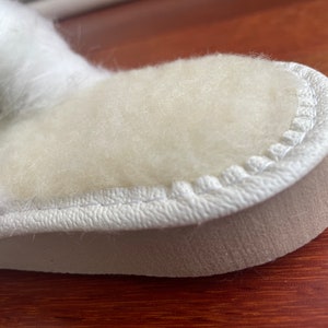 May include: Close-up of a white fuzzy slipper with a white stitched seam around the edge. The slipper has a beige sole.