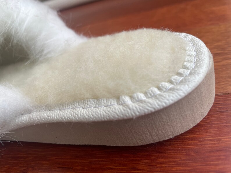 May include: Close-up of a white fuzzy slipper with a white stitched seam around the edge. The slipper has a beige sole.