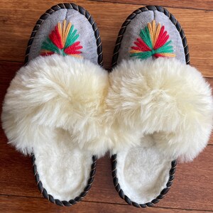 May include: A pair of gray sheepskin slippers with black trim and a colorful embroidered design. The slippers have a fluffy white lining.