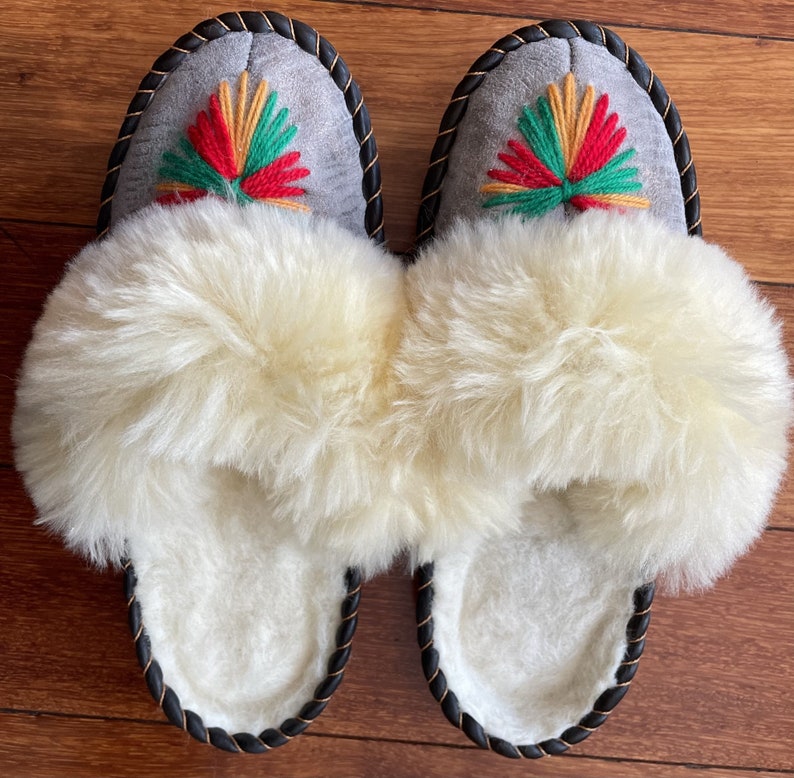 May include: A pair of gray sheepskin slippers with black trim and a colorful embroidered design. The slippers have a fluffy white lining.
