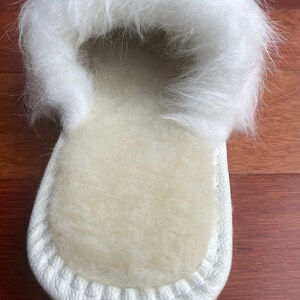 May include: White sheepskin slippers with a fluffy white fur trim. The slippers have a white stitched seam around the edge.