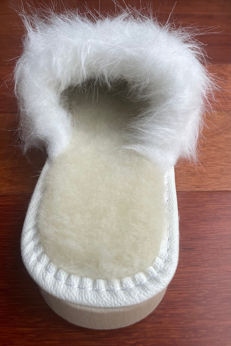 May include: White sheepskin slippers with a fluffy white fur trim. The slippers have a white stitched seam around the edge.